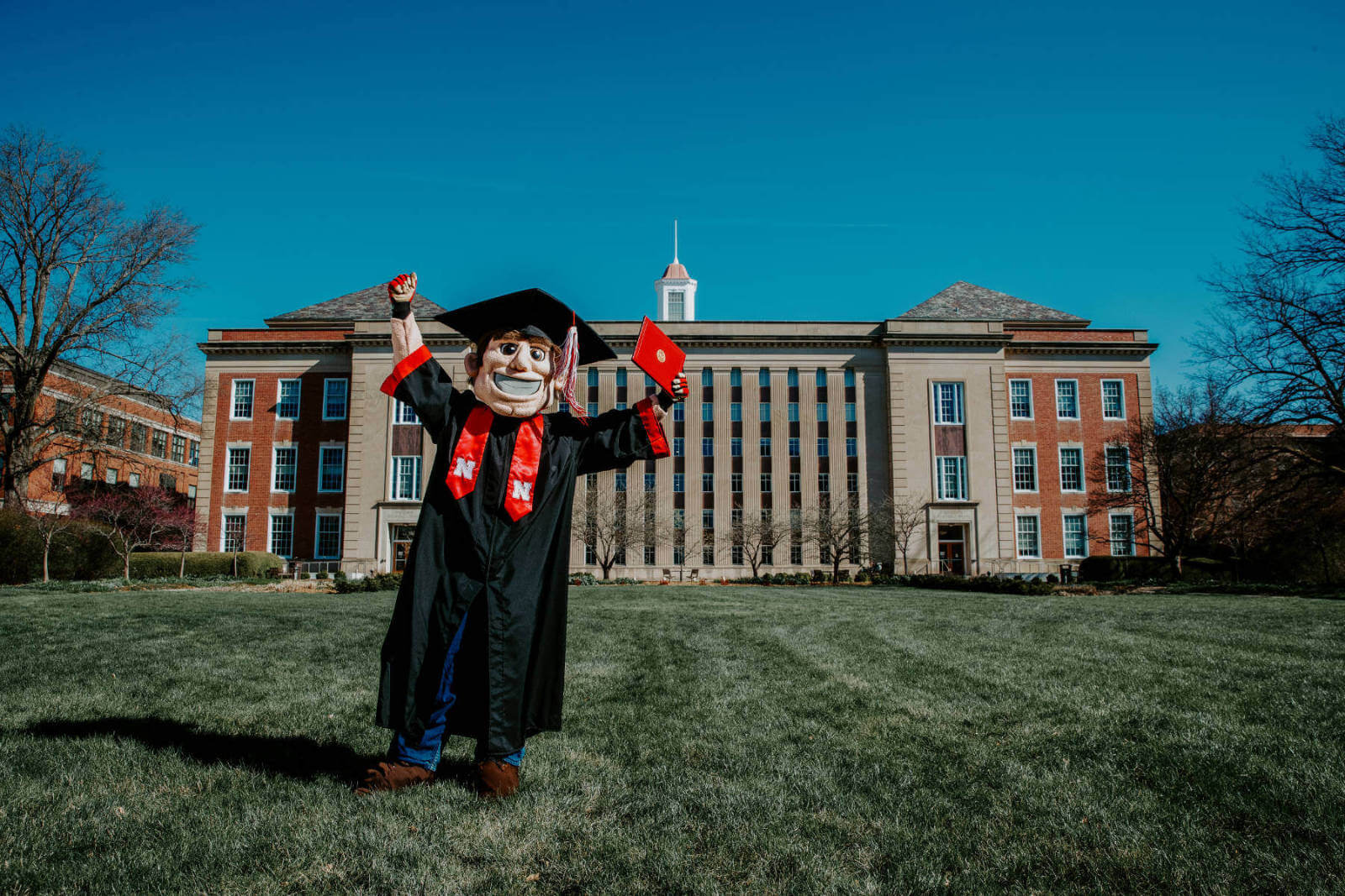 Herbie Husker wearing graduation gown and standing in front of Library