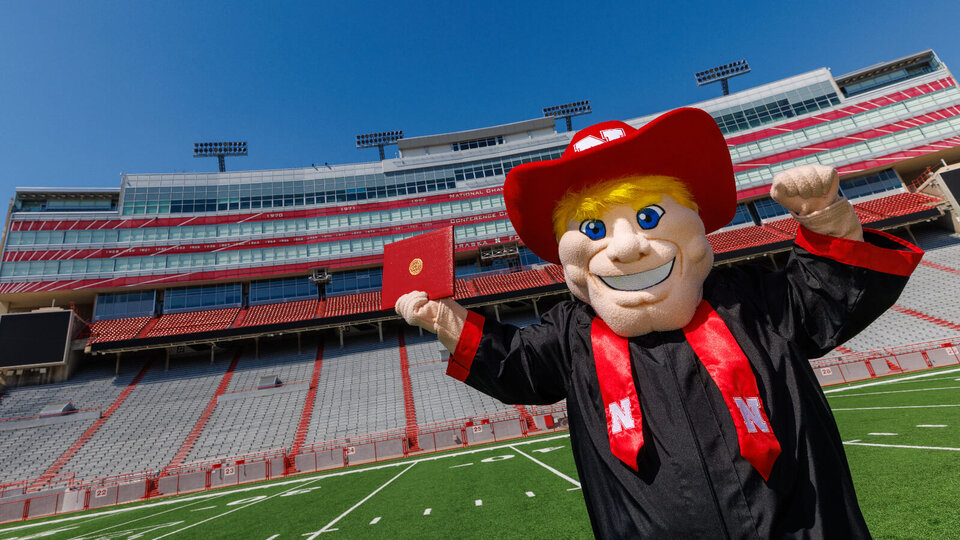 herbie husker holding a diploma and wearing graduation attire in memorial stadium