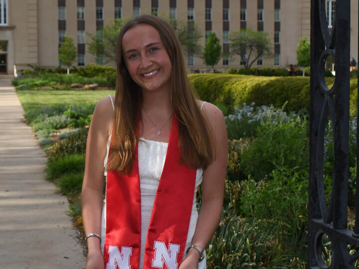 Graduate wearing a red sash with an "N" standing outdoors near a building and greenery.