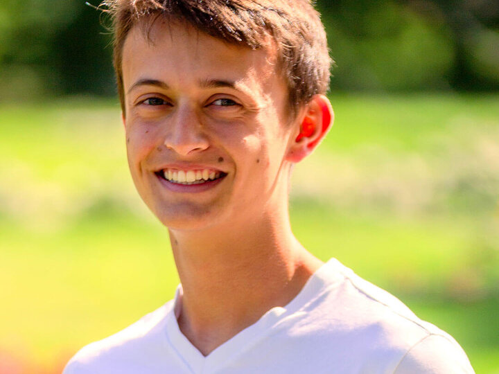 Smiling person outdoors with sunlight and greenery in the background.