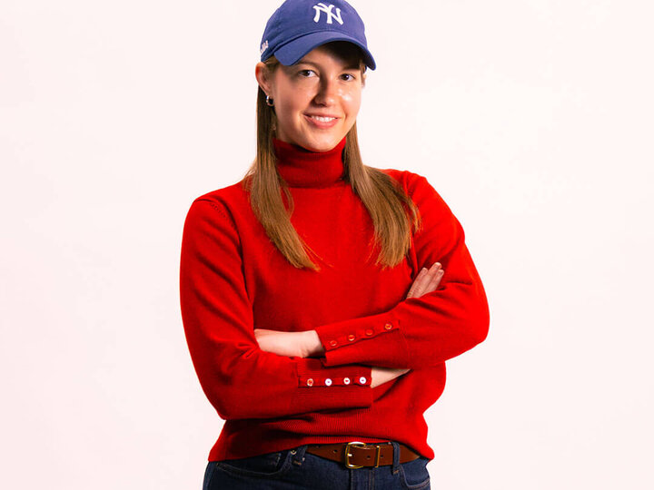 Person wearing a red sweater, blue cap, and jeans with arms crossed against a light background.